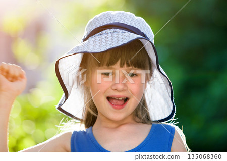 Close-up portrait of happy smiling little girl in a big hat. Child having fun time outdoors in summer. 135068360
