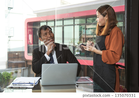 Asian businessman sitting at a modern cafe using a laptop while talking to a female waitress taking notes on a digital tablet, representing communication and customer service concept 135070331