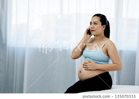 Pregnant Asian woman sitting on bed, holding her belly and talking on the phone with calm expression, representing motherhood, prenatal care, and healthy pregnancy lifestyle at home Pregnant Asian woman sitting on bed, holding her belly and talking on the phone with calm expression, representing motherhood, prenatal care, and healthy pregnancy lifestyle at home 135070333
