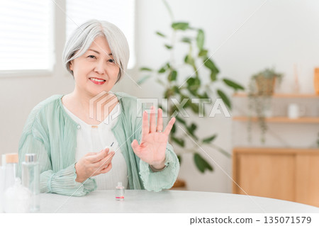 A senior woman with gray hair applies nail polish and nail oil to moisturize her nails. A senior woman with gray hair applies nail polish and nail oil to moisturize her nails. 135071579