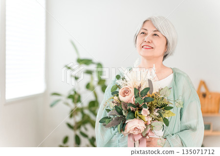 一位頭髮花白的老婦人手捧一束鮮花,在家中的客廳。 一位頭髮花白的老婦人手捧一束鮮花,在家中的客廳。 135071712