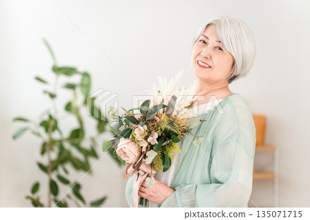 一位頭髮花白的老婦人在家裡的客廳裡手捧一束鮮花。 135071715
