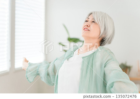 一位頭髮花白的老婦人在家裡的客廳裡深吸一口氣。 一位頭髮花白的老婦人在家裡的客廳裡深吸一口氣。 135072476