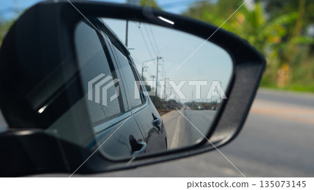 Car side mirror view reflecting the asphalt road and blue sky. Transportation, travel, and driving safety concept. Traffic conditions are clear. with electric pole side of road in Thailand. 135073145