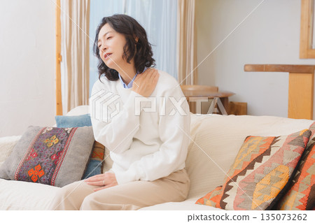 A middle-aged woman sitting on a sofa in the living room and massaging her shoulders 135073262