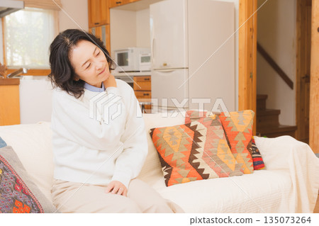 一位中年婦女坐在客廳的沙發上，正在按摩肩膀。 135073264