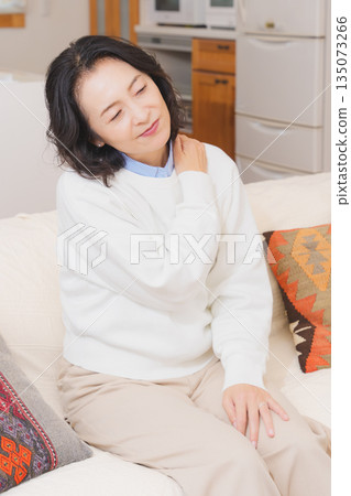 一位中年婦女坐在客廳的沙發上，正在按摩肩膀。 135073266