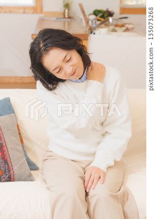 A middle-aged woman sitting on a sofa in the living room and massaging her shoulders 135073268