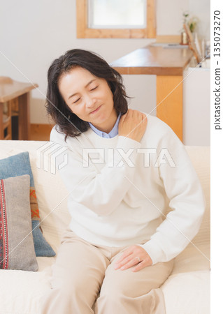 A middle-aged woman sitting on a sofa in the living room and massaging her shoulders A middle-aged woman sitting on a sofa in the living room and massaging her shoulders 135073270