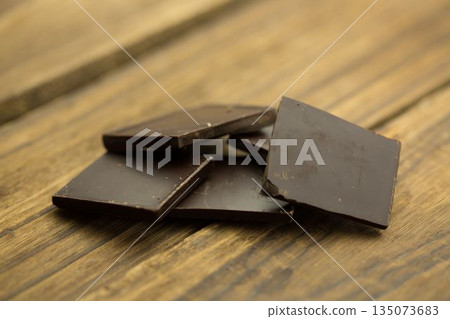 Dark chocolate squares are being stacked on rustic wooden tabletop, highlighting glossy surfaces Dark chocolate squares are being stacked on rustic wooden tabletop, highlighting glossy surfaces 135073683