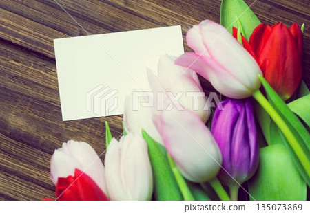 Flat design is featuring blank white card on rustic wooden tabletop with bright tulips lying beside 135073869