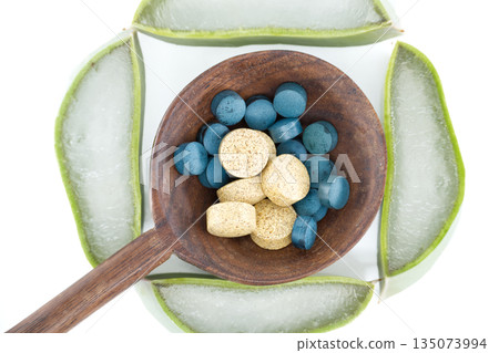Aloe vera slices arranged around a wooden spoon filled with blue and yellow pills 135073994