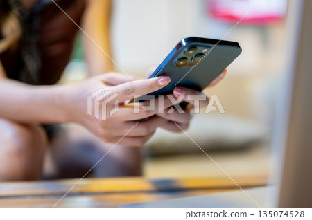 Close up of a woman's manicured hands holding phone sitting on sofa at wooden table in a living room Close up of a woman's manicured hands holding phone sitting on sofa at wooden table in a living room 135074528