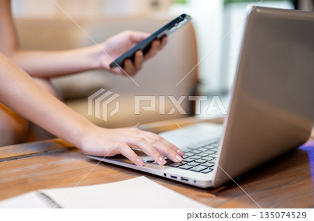 Close up woman hand holding phone aside laptop and notebook on table sitting at sofa in living room Close up woman hand holding phone aside laptop and notebook on table sitting at sofa in living room 135074529