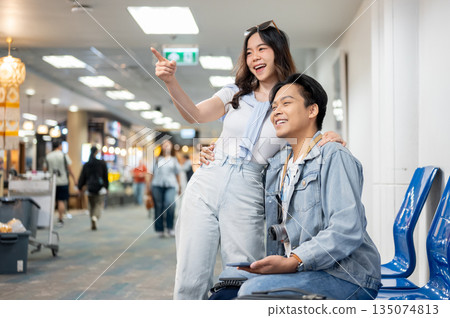 Asian tourist couple go on trip woman pointing aside sitting man as waiting for departure in airport Asian tourist couple go on trip woman pointing aside sitting man as waiting for departure in airport 135074813