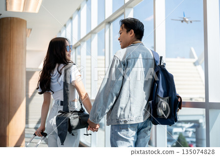 Back view of asian man and woman tourist couple go on a trip holding hands and walking in airport. 135074828