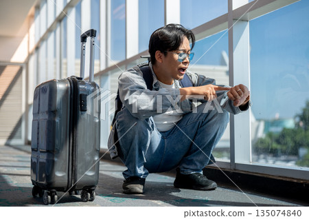 Frustrated asian man crouching looking at time on watch running late or missed a flight in airport Frustrated asian man crouching looking at time on watch running late or missed a flight in airport 135074840