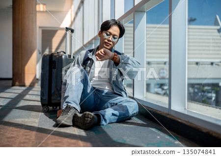 Asian man sitting on floor looking at time on watch waiting to depart or missed a flight in airport 135074841