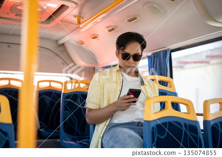 Asian man tourist wears shades holding looking at phone and sitting in shuttle bus while go on trip. Asian man tourist wears shades holding looking at phone and sitting in shuttle bus while go on trip. 135074855
