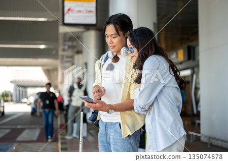 Asian tourist couple go on trip woman looking at man's phone as standing waiting for taxi in airport 135074878