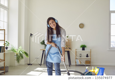 Housewife in earphones listening to music vacuuming the floor with vacuum cleaner at home. 135074977