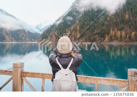 Happy woman traveler travel in jiuzhaigou national park in Sichuan, China. Tourist sightseeing and taking photo beautiful lake by mobile phone in Autumn season. Famous landmark near Chengdu, China 135075836