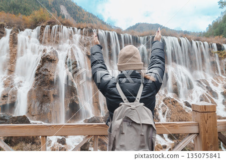 Happy woman traveler travel in jiuzhaigou national park in Sichuan, China. Tourist sightseeing Nuorilang Waterfall in Autumn season. Famous Landmark and Vacation near Chengdu, China Happy woman traveler travel in jiuzhaigou national park in Sichuan, China. Tourist sightseeing Nuorilang Waterfall in Autumn season. Famous Landmark and Vacation near Chengdu, China 135075841