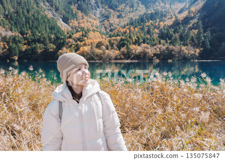 Happy woman traveler travel in jiuzhaigou national park in Sichuan, China. Tourist sightseeing Five Flower lake and snow mountain in Autumn season. Famous Landmark and Vacation near Chengdu, China Happy woman traveler travel in jiuzhaigou national park in Sichuan, China. Tourist sightseeing Five Flower lake and snow mountain in Autumn season. Famous Landmark and Vacation near Chengdu, China 135075847