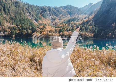 Happy woman traveler travel in jiuzhaigou national park in Sichuan, China. Tourist sightseeing Five Flower lake and snow mountain in Autumn season. Famous Landmark and Vacation near Chengdu, China 135075848