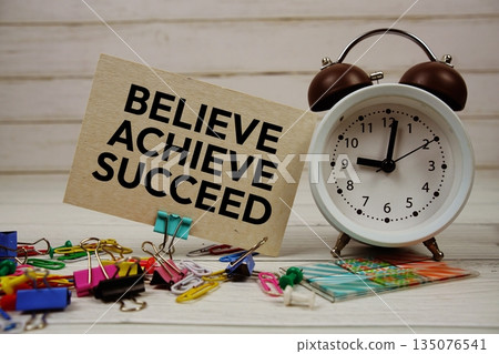 Believe, Achieve, Succeed text on paper card with alarm clock and stationary on wooden background Believe, Achieve, Succeed text on paper card with alarm clock and stationary on wooden background 135076541