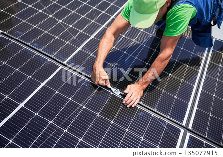 Worker building photovoltaic solar panel system on rooftop of house. Close up of man engineer installing solar module with help of wrench outdoors. Alternative and renewable energy generation concept. Worker building photovoltaic solar panel system on rooftop of house. Close up of man engineer installing solar module with help of wrench outdoors. Alternative and renewable energy generation concept. 135077915