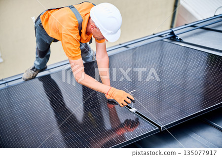 Worker building photovoltaic solar panel system on rooftop of house. Man engineer in helmets and gloves installing solar module with help of hex key outdoors. Alternative energy generation concept 135077917