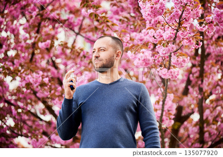 Man allergic using medical nasal drops, suffering from seasonal allergy at spring in blossoming garden. Handsome man showing nasal spray near blooming tree outdoors. Spring allergy concept. 135077920