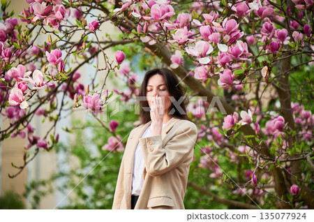Woman allergic using medical nasal drops, suffering from seasonal allergy at spring in blossoming garden. Young woman treating runny nose in front of blooming tree outdoors. Spring allergy concept. Woman allergic using medical nasal drops, suffering from seasonal allergy at spring in blossoming garden. Young woman treating runny nose in front of blooming tree outdoors. Spring allergy concept. 135077924