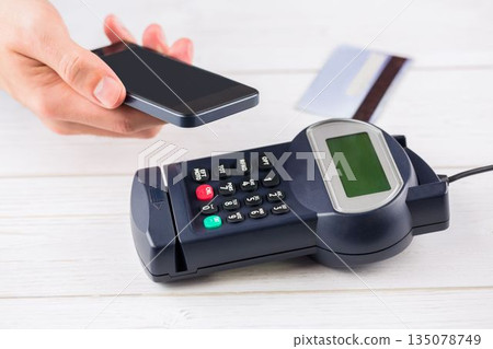 Right hand is holding smartphone over point-of-sale terminal on white wooden tabletop, flat design Right hand is holding smartphone over point-of-sale terminal on white wooden tabletop, flat design 135078749
