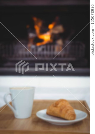 Coffee cup by croissant in plate on table against fireplace 135078956