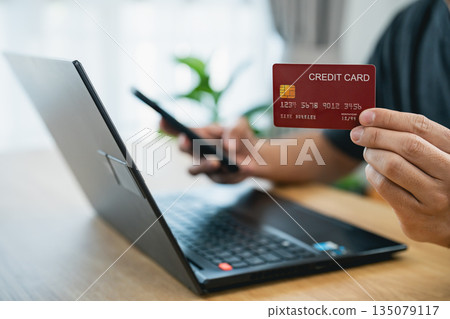 Online shopping and credit card payment via mobile and laptop. Man holding a credit card buying items online, making financial e-commerce transactions, internet banking at home, convenient and secure. 135079117