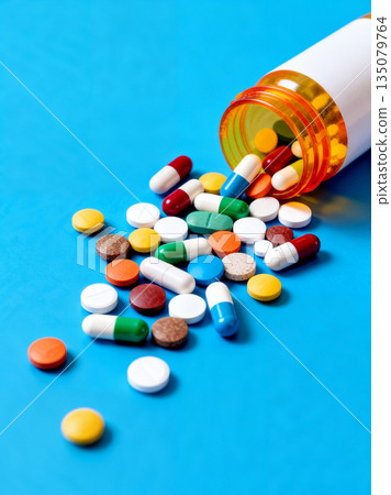 Assorted colorful prescription pills and capsules spilling out of an amber plastic bottle on a blue background. Medical healthcare, pharmacy, and pharmaceutical drug treatment concept. 135079764