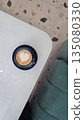 Overhead view of a heart shaped latte art in a ceramic cup on a modern white table with terrazzo flooring and soft seating.  135080330