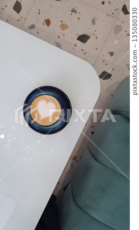 Overhead view of a heart shaped latte art in a ceramic cup on a modern white table with terrazzo flooring and soft seating. Overhead view of a heart shaped latte art in a ceramic cup on a modern white table with terrazzo flooring and soft seating. 135080330