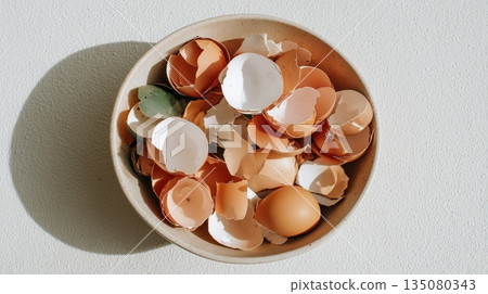 Natural brown and white eggshells collected in a wooden bowl, placed under soft sunlight on a textured white surface.  135080343