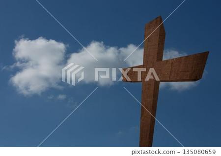 Rough-hewn wooden cross is standing against clear blue sky with drifting white cloud in flat design 135080659