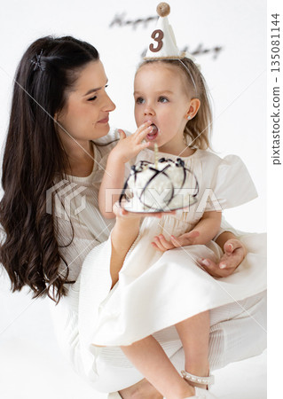A mother and her young daughter celebrate a birthday together, with the little girl eating cake. A mother and her young daughter celebrate a birthday together, with the little girl eating cake. 135081144