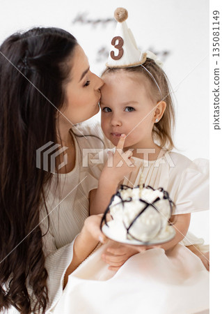 A mother kisses her daughter's forehead as they celebrate her third birthday, holding a cake. 135081149