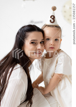 A mother and her young daughter are embracing, celebrating the daughter's third birthday with a festive hat and smiles. A mother and her young daughter are embracing, celebrating the daughter's third birthday with a festive hat and smiles. 135081157