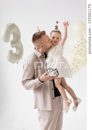 A father kisses his daughter on the cheek while holding her at her third birthday celebration, complete with a cake and balloons. A father kisses his daughter on the cheek while holding her at her third birthday celebration, complete with a cake and balloons. 135081179