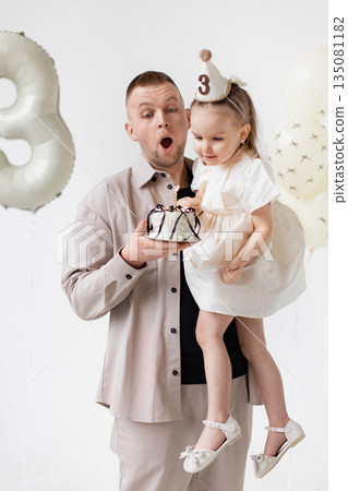 A father holds his daughter, celebrating her third birthday with a cake and balloons. 135081182