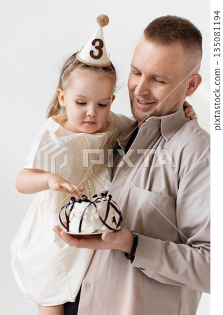 A father holds his daughter, who is wearing a party hat, as they celebrate her third birthday with a cake. A father holds his daughter, who is wearing a party hat, as they celebrate her third birthday with a cake. 135081194