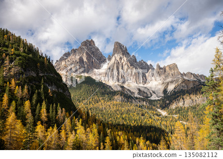 Dramatic rocky Dolomites mountain peaks above autumn forest under blue sky Dramatic rocky Dolomites mountain peaks above autumn forest under blue sky 135082112