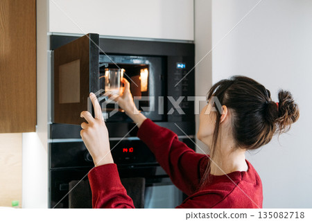 Woman reaches into microwave to heat glass of milk in modern kitchen. Heating oat milk, vegan latte preparation, dairy free lifestyle. Woman reaches into microwave to heat glass of milk in modern kitchen. Heating oat milk, vegan latte preparation, dairy free lifestyle. 135082718
